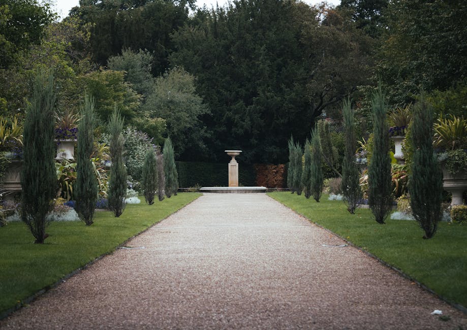 A symmetrical garden pathway lined with tall, narrow cypress trees on both sides, leading to a fountain or decorative planter at the far end. The pathway is composed of small, light-colored gravel, bo