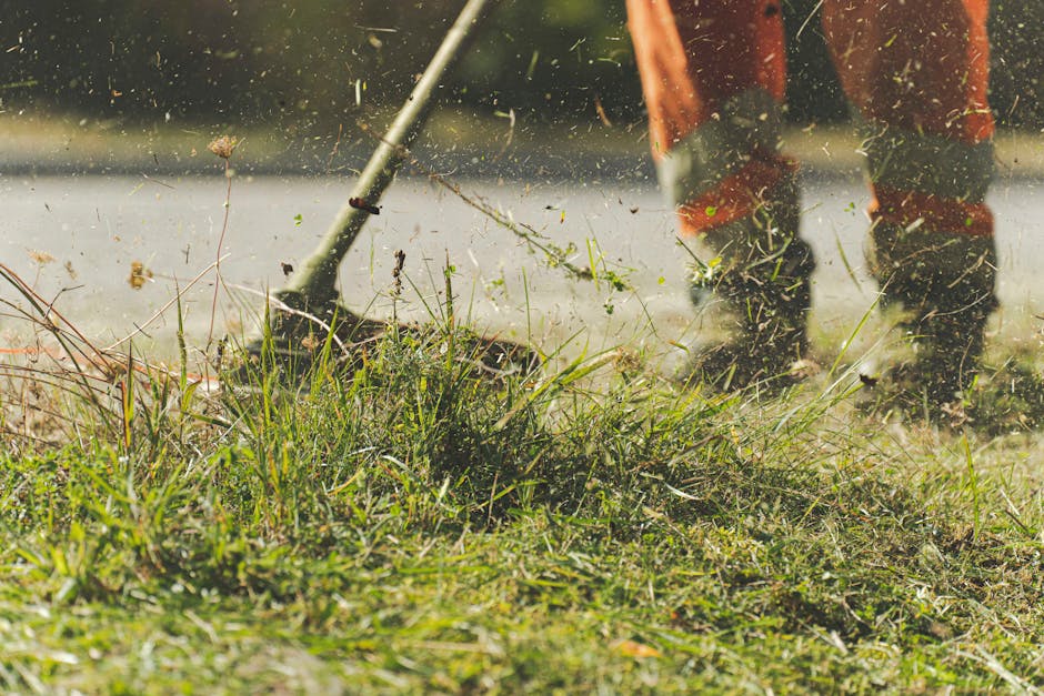 Close-up view of a garden lawn being maintained by a gardener using a powered tiller, with the gardener's legs and boots visible in the background. The soil and grassy plants are being disturbed, crea